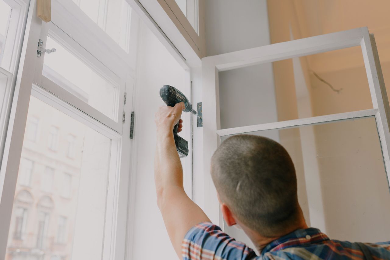 Features Back view of unrecognizable craftsman using cordless screwdriver and assembling window while screwing metal hinge during renovation works in apartment