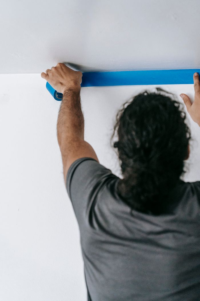 Features Adult applying blue tape to ceiling for painting, focused on precision.
